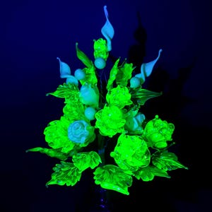 Peut inclure: Un arrangement floral en verre composé de plusieurs roses et de fleurs en forme de lys calla qui émettent une lueur vert néon brillante sous une lumière ultraviolette, entremêlées de feuilles en verre détaillées et de petits accents sphériques.