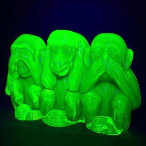 May include: A fluorescent green glass figurine depicting the three wise monkeys. The sculpture features one monkey covering its eyes, one covering its ears, and one covering its mouth, all glowing intensely against a dark background under UV light.