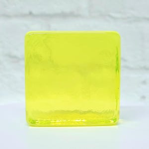 Puede incluir: Un objeto cuadrado de vidrio transparente en un tono amarillo vibrante. El vidrio tiene una superficie ligeramente texturizada y los bordes son redondeados. El objeto est&aacute; sobre un fondo blanco, con un toque de pared de ladrillo al fondo.