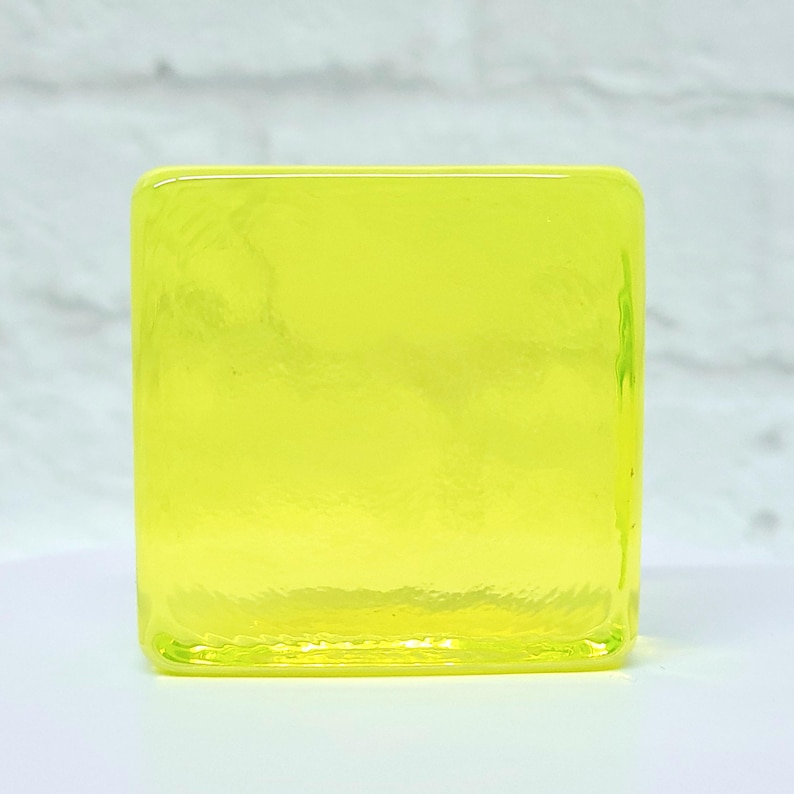 Puede incluir: Un objeto cuadrado de vidrio transparente en un tono amarillo vibrante. El vidrio tiene una superficie ligeramente texturizada y los bordes son redondeados. El objeto est&aacute; sobre un fondo blanco, con un toque de pared de ladrillo al fondo.