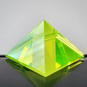 May include: A bright green glass pyramid with a smooth, polished surface. The pyramid is made of clear glass with a green tint, and it reflects light beautifully.