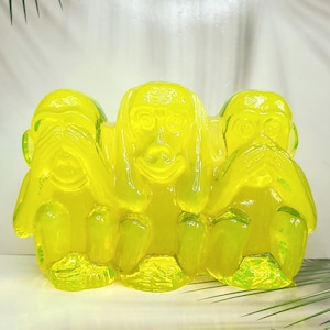 May include: A translucent neon yellow glass sculpture depicting three monkeys sitting side by side. The first monkey covers its eyes, the second monkey covers its ears, and the third monkey covers its mouth, representing the three wise monkeys motif.