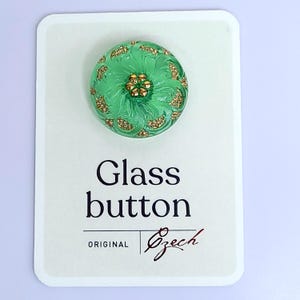 May include: A round, green glass button with a floral design and gold accents. The button is on a white card with the text "Glass button" and "Original Czech".