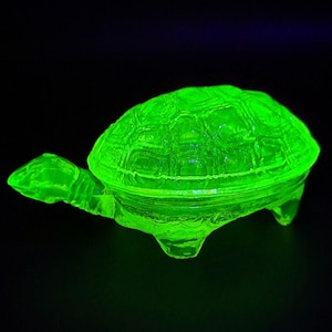 May include: A bright green glass turtle trinket box with a detailed shell design. The box is made of uranium glass and glows under a black light.