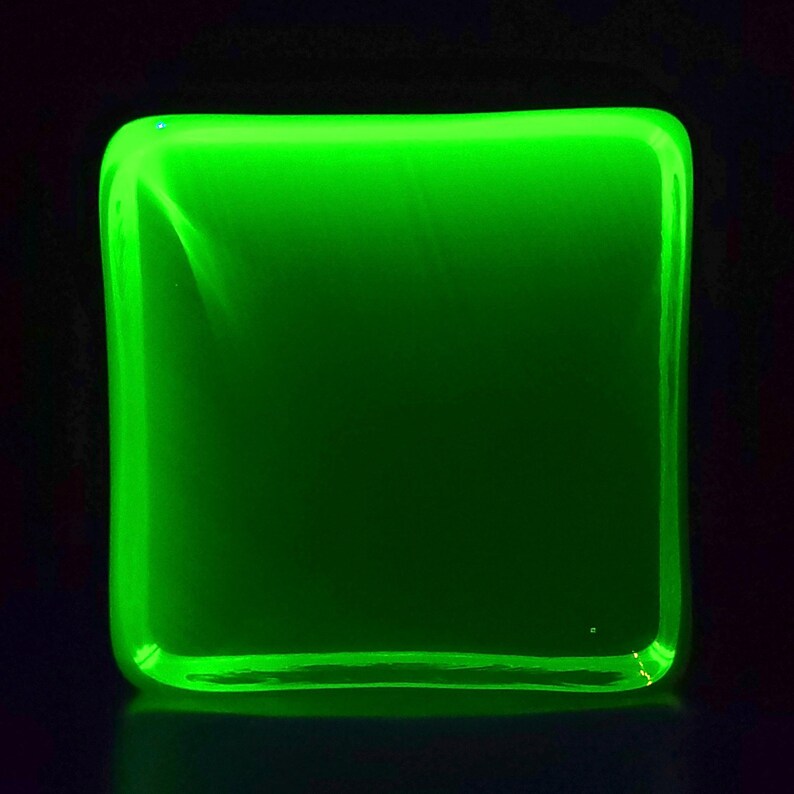 Puede incluir: Un objeto cuadrado brillante con esquinas redondeadas, que emite una luz verde vibrante. El objeto parece estar hecho de un material transl&uacute;cido, con una superficie lisa y un ligero reflejo en la base.