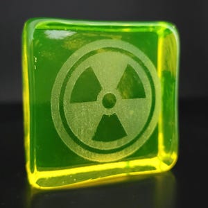 Op de afbeelding: Een vierkant, transparant, groen glazen blok met een gele rand. Het blok heeft een geëtst stralingssymbool in het midden. Het glas heeft een lichte gloed en de achtergrond is donker.