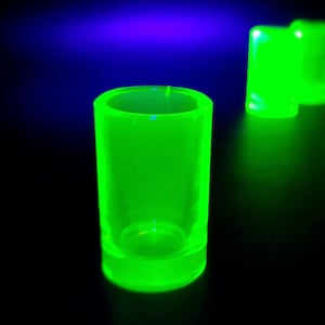 Può includere: Primo piano di tre bicchierini verde neon. I bicchierini, cilindrici e con base spessa, brillano intensamente su uno sfondo scuro. Uno è a fuoco, gli altri due sfocati sullo sfondo, creando profondità.