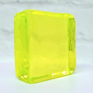 Puede incluir: Un bloque de vidrio amarillo ne&oacute;n transl&uacute;cido con bordes redondeados. El bloque rectangular tiene una superficie lisa y brillante. El vidrio parece s&oacute;lido y de color uniforme, con una ligera textura visible en los lados. Mide unos 5 cm de alto.