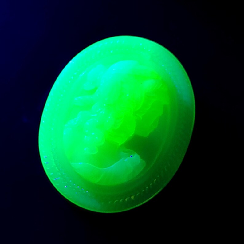May include: A glowing green, oval-shaped cameo with a profile of a woman. The cameo is set against a black background. The cameo is made of a translucent material and has a detailed relief carving.
