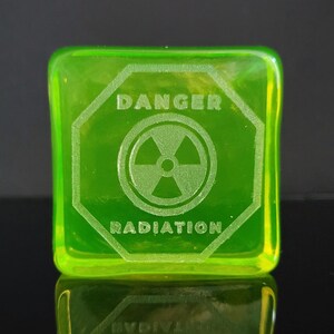 Op de afbeelding: Een vierkant, heldergroen glazen blok met de woorden "DANGER" en "RADIATION" in het oppervlak gegraveerd. Een stralingssymbool staat in het midden. Het blok is licht transparant, met afgeronde randen.