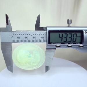 May include: A light green, oval-shaped cameo is measured with a digital caliper. The cameo features a detailed relief carving of a classical scene. The caliper displays a measurement of 43.30 mm.