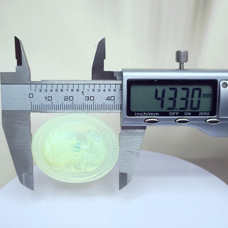 May include: A light green, oval-shaped cameo is measured with a digital caliper. The cameo features a detailed relief carving of a classical scene. The caliper displays a measurement of 43.30 mm.