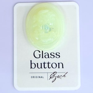 May include: A pale yellow glass button with an oval shape, featuring a detailed relief design within a rope-like border. The button is on a white card with the words "Glass button" and "ORIGINAL Czech" printed in black.