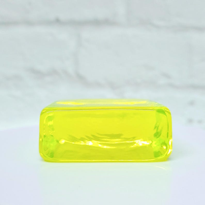 Puede incluir: Un objeto rectangular de color amarillo ne&oacute;n con esquinas redondeadas. El material transl&uacute;cido permite el paso de la luz, creando un efecto brillante. El objeto est&aacute; sobre una superficie blanca y una pared de ladrillos blanca borrosa.