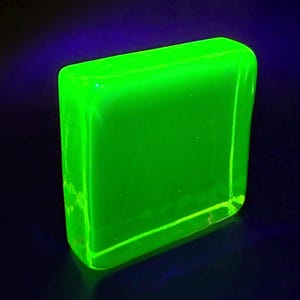 Op de afbeelding: Een lichtgevend, rechthoekig blok met afgeronde hoeken, dat een levendige neon groene kleur uitstraalt. Het object lijkt doorschijnend te zijn, met een glad oppervlak. De achtergrond is diepblauw, wat een sterk contrast vormt met het heldere groen.