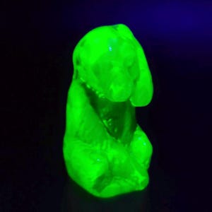 May include: A glowing green figurine of a dog, possibly a beagle, with long floppy ears and a collar. The sculpture is illuminated with a bright green light against a dark background, highlighting its form and details.