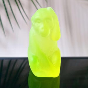 May include: A neon green monkey figurine. The sculpture is smooth and has a glossy finish. The monkey is sitting upright with its arms at its sides and its ears perked up. The figurine is reflected on a black surface.