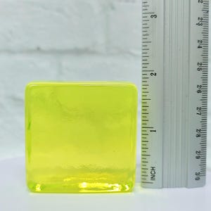 Puede incluir: Un bloque de vidrio amarillo ne&oacute;n transl&uacute;cido de forma cuadrada. Una regla est&aacute; colocada junto al bloque, mostrando medidas en pulgadas. El bloque tiene bordes redondeados y una superficie lisa.