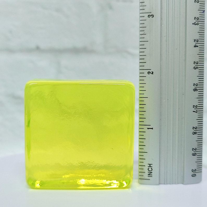 Puede incluir: Un bloque de vidrio amarillo ne&oacute;n transl&uacute;cido de forma cuadrada. Una regla est&aacute; colocada junto al bloque, mostrando medidas en pulgadas. El bloque tiene bordes redondeados y una superficie lisa.