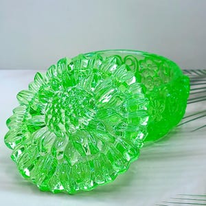 Op de afbeelding: Een levendig neongroen glazen sieradendoosje met een afneembaar deksel. Het deksel heeft een gedetailleerd, verhoogd zonnebloemachtig bloemmotief met gestructureerde bloemblaadjes. De basis van de houder toont een herhalend reliëf bloemmotief.