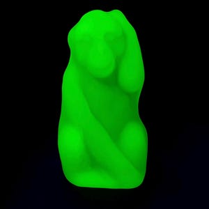 May include: A bright green sculpture of a seated figure with its hand raised to its head. The sculpture has smooth, rounded features and is set against a black background. The figure appears to be in a pose of contemplation or distress.