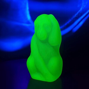 May include: A bright green figurine of a seated dog with its head resting on its front paws. The sculpture is illuminated against a dark blue background, creating a striking contrast. The dog's form is smooth and rounded, with a slightly abstract appearance.