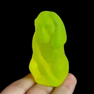May include: A translucent, neon yellow gummy candy shaped like a monkey. The figure features a smooth, molded surface with defined facial features, including ears and a snout, and a curved body posture. The candy has a vibrant, semi-transparent appearance.