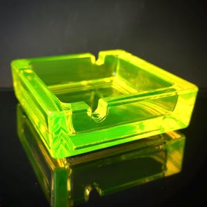 Può includere: Un posacenere quadrato verde neon in vetro trasparente. Il posacenere ha una forma rettangolare con una piccola scanalatura su ogni lato per appoggiare le sigarette. Il posacenere è appoggiato su una superficie nera riflettente.