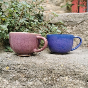 Peut inclure: Deux tasses en céramique, l'une de couleur violet profond et l'autre bleu vif. Les deux tasses ont une texture mouchetée et des formes arrondies avec de grandes anses. Les tasses sont posées sur une surface en pierre, avec de la verdure en arrière-plan.