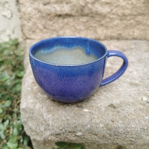 Peut inclure: Une tasse en céramique faite à la main avec un glaçage bleu profond. La tasse a une forme arrondie avec une anse et un intérieur bleu plus clair. La tasse est posée sur une surface en pierre.