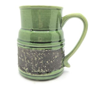 Peut inclure: Une tasse en céramique faite à la main avec un dessus et une anse émaillés verts. La partie inférieure présente une bande texturée brun foncé avec des détails mouchetés. La tasse est conçue pour les boissons chaudes et a une capacité d'environ 350 ml.