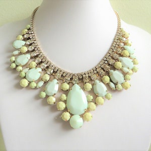 May include: A statement necklace featuring a gold-tone chain and a bib of large, light green and yellow teardrop-shaped and round gemstones. The necklace is adorned with clear, sparkling rhinestones.