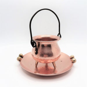 May include: A small copper cauldron with a black handle, resting on a matching copper plate. The cauldron has three small legs and a hammered texture. The plate has two decorative gold-colored spheres.