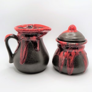 May include: A red and black ceramic creamer and sugar set. The creamer has a handle and a rounded body. The sugar bowl has a lid and a rounded body. Both pieces have a drip glaze design.