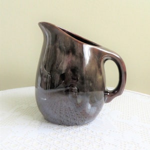 May include: A brown ceramic pitcher with a glossy finish. The pitcher has a wide mouth and a handle on the side.