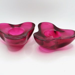May include: Two pink glass candle holders shaped like a heart. The holders are made of clear glass with a pink tint.