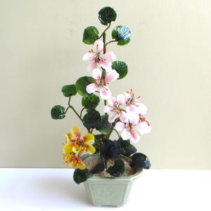 Peut inclure: Composition florale décorative dans un pot carré vert clair. L'arrangement présente des fleurs roses et jaunes avec des feuilles et des tiges vertes. Le pot est rempli d'un matériau clair, peut-être du sable ou de petites pierres.