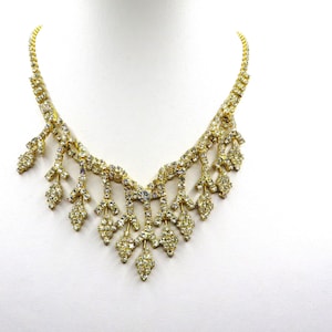 May include: A gold necklace with a delicate, cascading design featuring clear rhinestones. The necklace has a V-shaped neckline and is adorned with small, sparkling stones.
