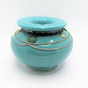 May include: A turquoise ceramic ashtray with a silver rim and decorative green and gold accents.