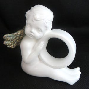 Pu&ograve; includere: Statuetta di angelo in ceramica bianca con anello. L'angelo ha lineamenti dettagliati, capelli ricci e ali dorate e iridescenti. La statuetta &egrave; seduta e tiene l'anello.