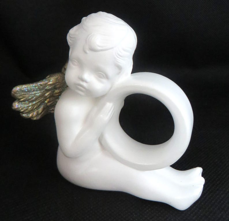 Pu&ograve; includere: Statuetta di angelo in ceramica bianca con anello. L'angelo ha lineamenti dettagliati, capelli ricci e ali dorate e iridescenti. La statuetta &egrave; seduta e tiene l'anello.