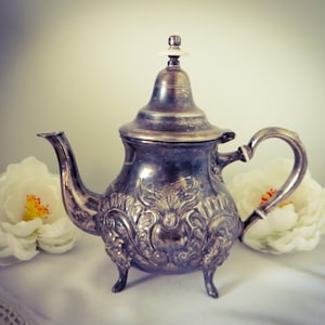 May include: Ornate silver teapot with a curved handle and spout, resting on three small feet. The teapot features detailed floral and scrollwork patterns. Two large white flowers with yellow centers flank the teapot, set against a soft white background.