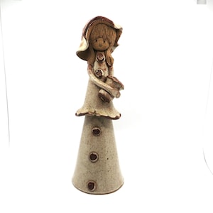 May include: A tall, speckled ceramic figurine of a person wearing a wide-brimmed hat and a dress. The figure has brown hair, a small mouth, and holds a small pot. The base is a wide, conical shape with three circular brown accents.