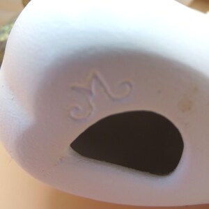 Pu&ograve; includere: Primo piano di un oggetto in ceramica bianca con un'apertura scura. L'oggetto ha una superficie liscia e una "M" decorativa in corsivo incisa sul lato. L'oggetto sembra essere un pezzo decorativo o parte di un progetto artistico pi&ugrave; ampio.