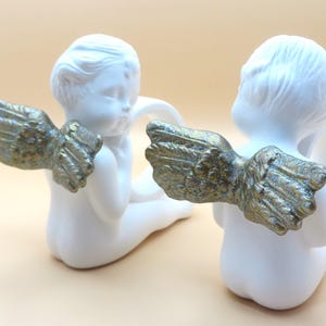 Pu&ograve; includere: Due statuette di cherubini in ceramica bianca con ali color oro. I cherubini sono in posizione seduta, con la schiena rivolta verso l'osservatore. Le ali sono dettagliate e strutturate, aggiungendo un tocco di eleganza.