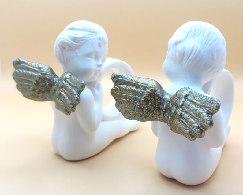 Pu&ograve; includere: Due statuette di cherubini in ceramica bianca con ali color oro. I cherubini sono in posizione seduta, con la schiena rivolta verso l'osservatore. Le ali sono dettagliate e strutturate, aggiungendo un tocco di eleganza.