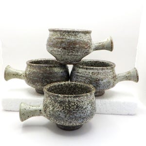 Peut inclure: Quatre tasses en céramique faites à la main avec des poignées. Les tasses sont gris et marron mouchetés avec un aspect rustique et texturé. Les tasses sont empilées les unes sur les autres, mettant en valeur leur design et leur savoir-faire uniques.