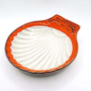 May include: A white ceramic seashell-shaped dish with an orange rim. The dish has a raised design on the rim and is perfect for serving small snacks or sauces.