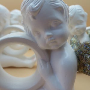 Pu&ograve; includere: Una collezione di statuette di cherubini bianchi, ognuna con un'espressione serena e lineamenti delicati. Un cherubino ha un'ala dorata scintillante. Le statuette sono disposte in gruppo, suggerendo un tema decorativo.