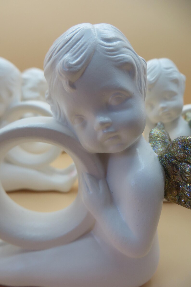 Pu&ograve; includere: Una collezione di statuette di cherubini bianchi, ognuna con un'espressione serena e lineamenti delicati. Un cherubino ha un'ala dorata scintillante. Le statuette sono disposte in gruppo, suggerendo un tema decorativo.